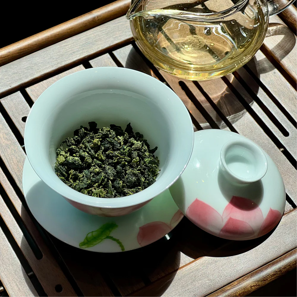 Tieguanyin oolong tea leaves in a white gaiwan on a wooden tea tray, with a glass fairness pitcher of golden tea liquor