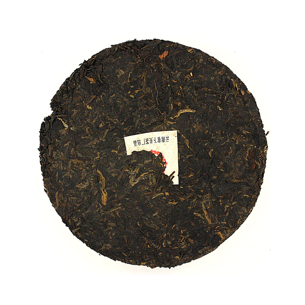 A dark compressed pu'er tea cake with a small paper label in the centre, showing tightly pressed aged tea leaves